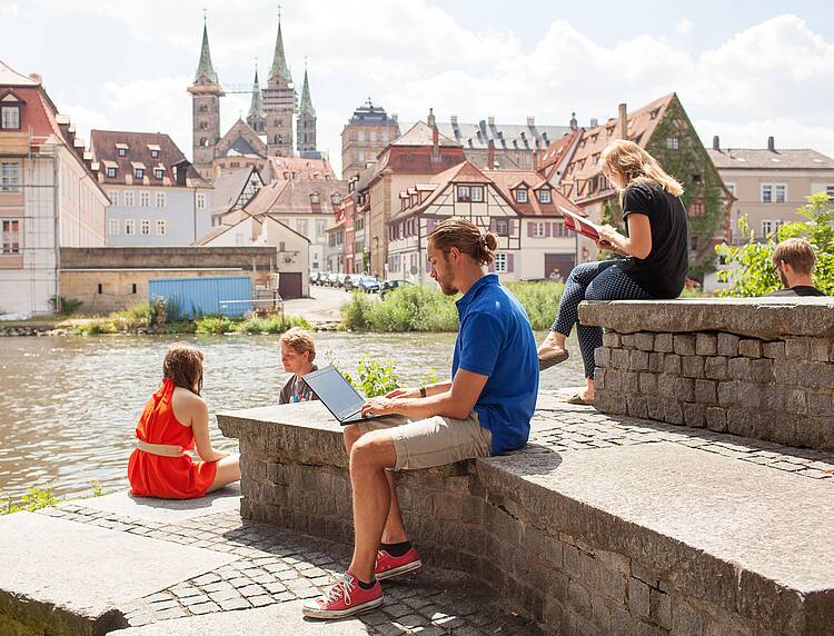 Universitätsstadt Bamberg (Foto: Universität Bamberg/Tim Kipphahn) Foto: Studierende sitzen am Ufer des Mains, im Hintergrund Bamberg